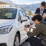 空港のレンタカー受取場所で車体の傷をスマートフォン撮影する旅行者と係員